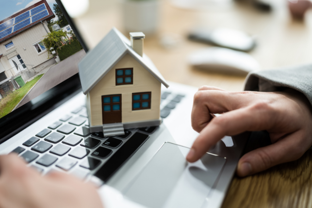 Close-up of hands using a laptop touchpad with a small house model, symbolizing how to register a rental property online.