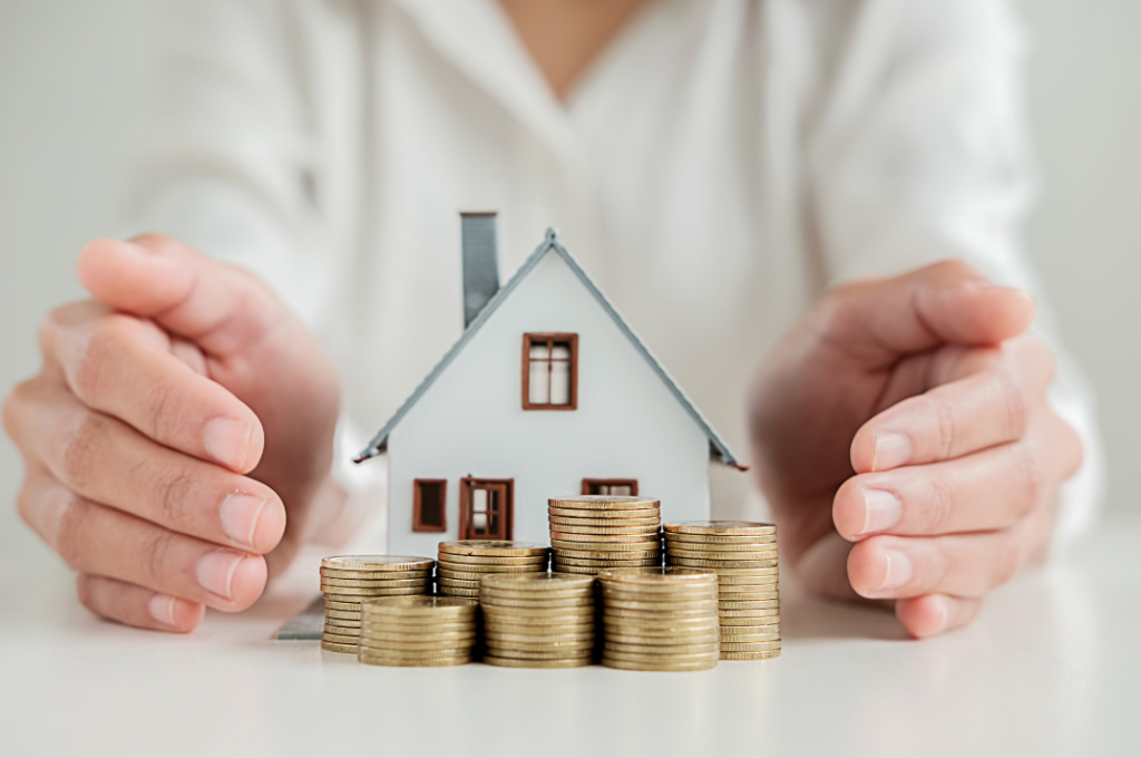 Hands protectively placed around a small house model with stacked coins in front, illustrating the concept of listing rental properties for financial gain.
